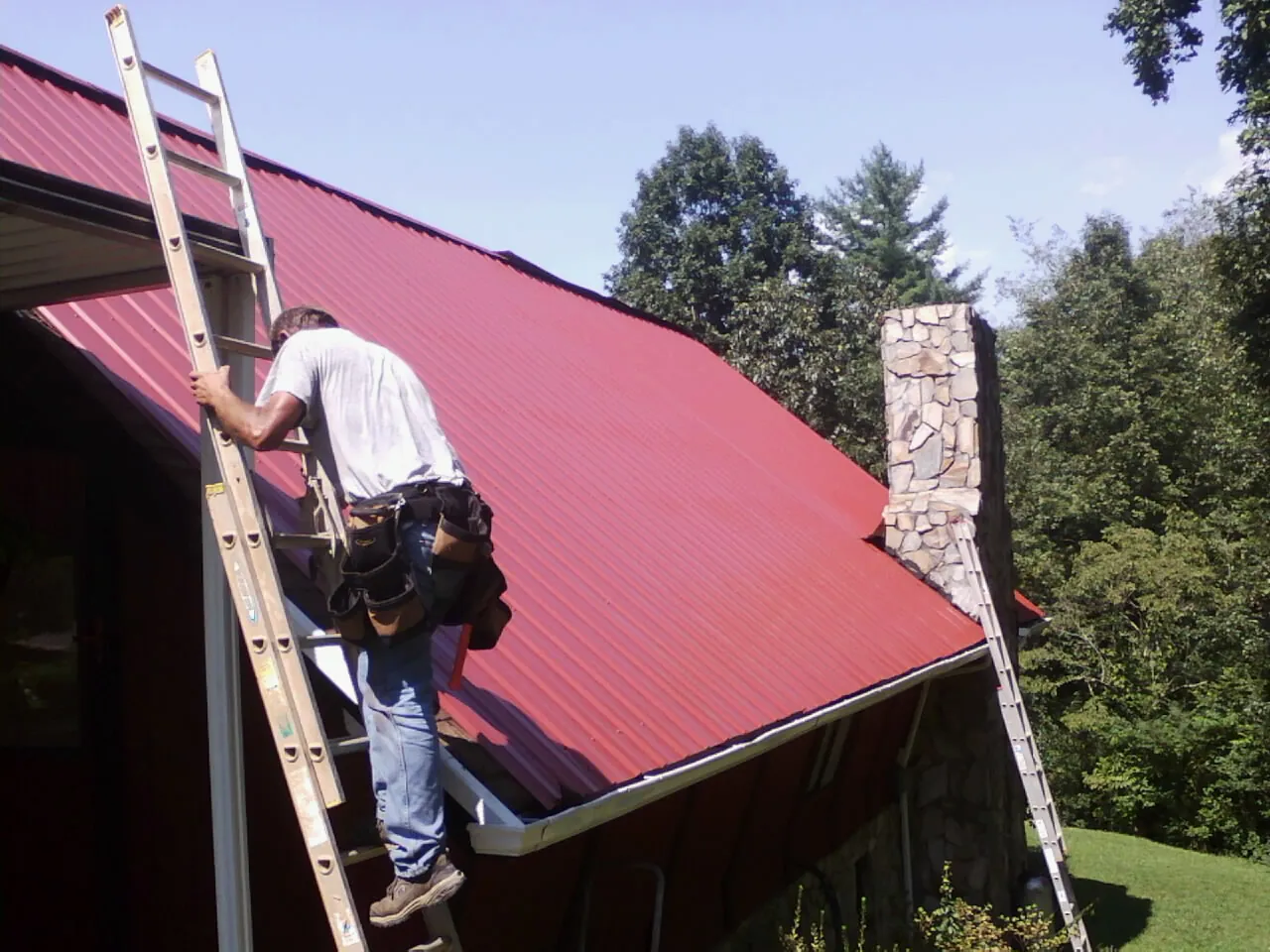 Roofer on a ladder beside a red metal roof and stone chimney — Soffit & Fascia Repair by Roofing Brothers in Bremen